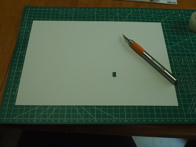 A letter-sized sheet of card-stock with a small rectangle cut out. The
sheet is sitting on a cutting mat and has an exacto knife sitting on
it.